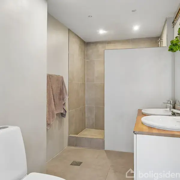 Toilet står ved siden af en åben bruseniche med beige fliser. På modsatte side er en dobbeltvask på et træbord, hvori der er planter og spejl ovenover.