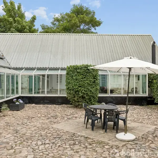 Et havebord med stole står under en parasol på en brostensbelagt terrasse. En bygning med store vinduer, omkranset af grønne hække og træer, udgør baggrunden.