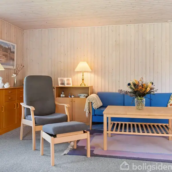 En hyggelig stue med en grå lænestol og fodskammel, en blå sofa, trægulve, reoler, bord med blomster i en vase, og lamper i blødt lys.