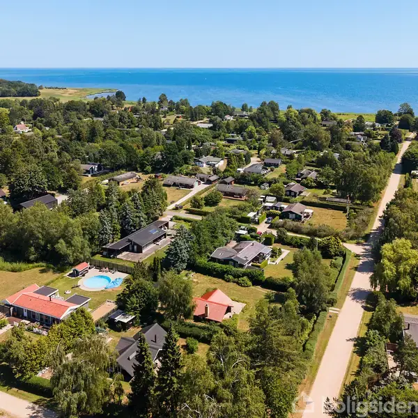 Dronebillede af en kystlandsby med spredte huse og grønne haver i et frodigt landskab tæt på havet. Veje snor sig gennem området, mens havet strækker sig mod horisonten.