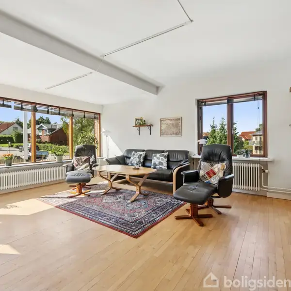 Stue med lædersofa og lænestole omkring et sofabord på et tæppe. Store vinduer med udsigt til en have, vægur og vinreol ved siden af.