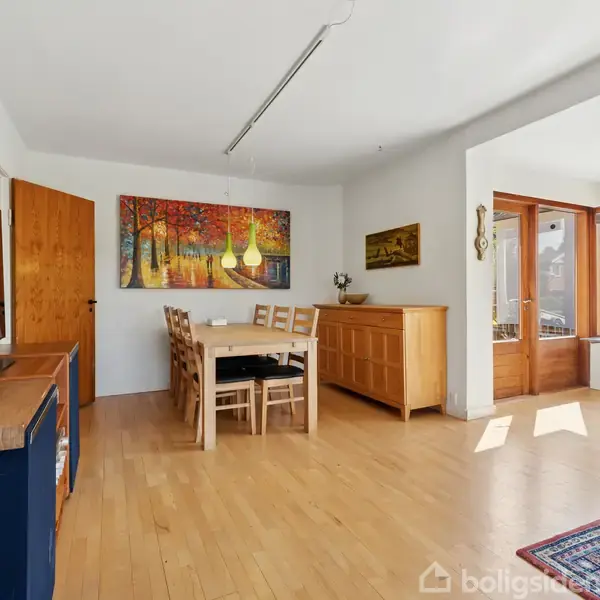 Et lyst spisestueområde med et træbord og seks stole. Et maleri af en skov hænger på væggen. Store vinduer lukker lys ind i rummet. En tæppe ligger på trægulvet.