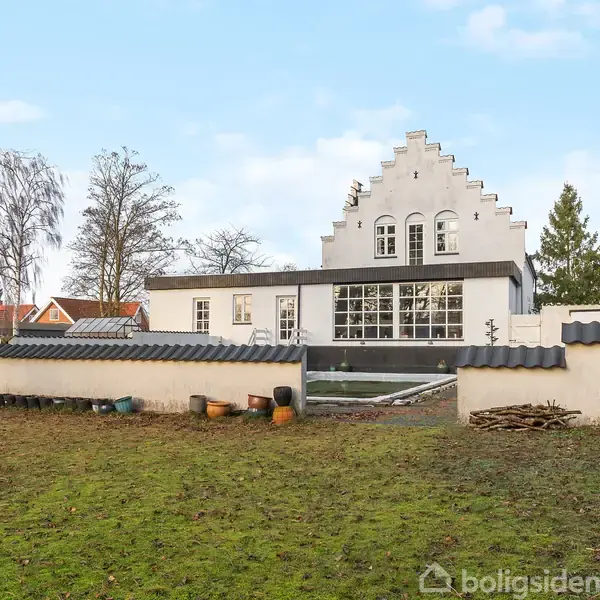 Hvid bygning med trappegavl står på en græsplæne omkranset af lav mur. Træer ses i baggrunden under en klar himmel.