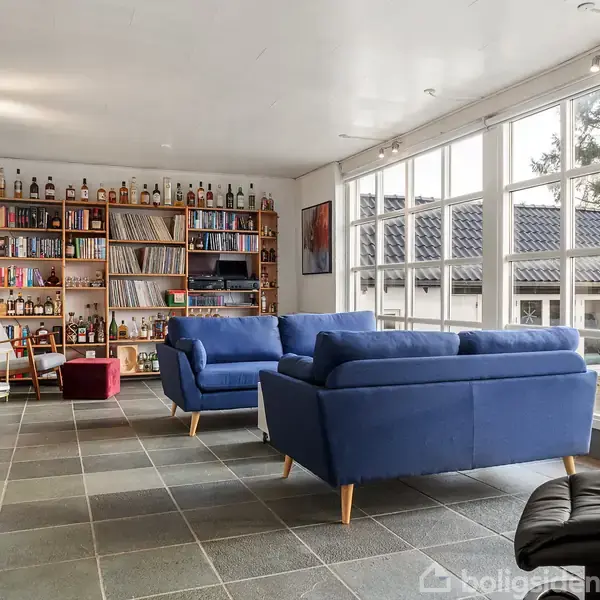 To blå sofaer står i en lys stue med store vinduer og gråt flisegulv. En reol fyldt med bøger, spiritusflasker og plader står mod væggen.