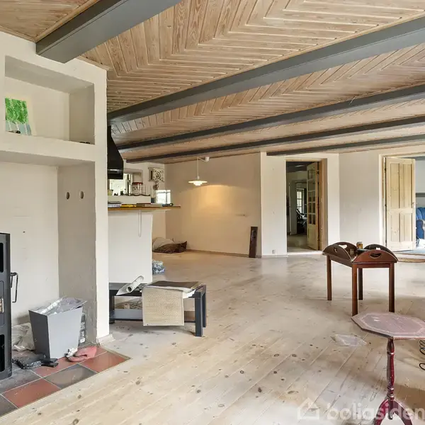 Brændeovn opvarmer stue med trægulv og synlige bjælker. Et lille bord og stole står nær en gulvlampe, mens der er nogle åbne kasser og en fodbold omkring.