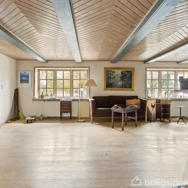 En stue med trægulv og træloft har en mørk sofa, skrivebord, og lamper. Værelserne oplyses af vinduer, og der er billeder på væggene samt diverse dekorative genstande.