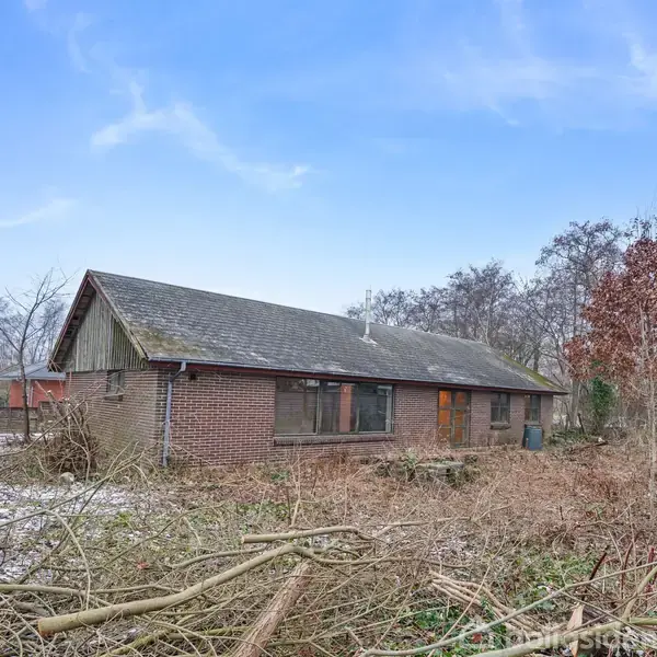 En murstensbygning står uberørt, omgiven af vilde buske og træer i en vinterlig have. En blå himmel med spredte skyer danner baggrunden.