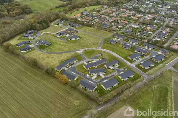 En gruppe moderne huse er placeret i et grønt område omgivet af træer og marker. De danner et nabolag med indre veje, der forbinder til en nærliggende bydel.