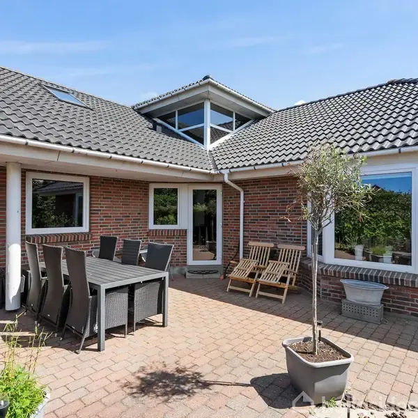 Terrasse med spisebord og stole placeret på fliser. To træbænke står ved siden af en murstensvæg. En stor, grå husmur med flere vinduer omkranser området. Blå himmel ses i baggrunden.