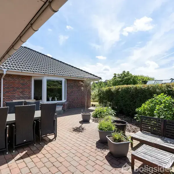 Terrassemøbler står på en brolagt terrasse foran et rødt murstenshus med hvide vinduesrammer. Krukker med planter er placeret rundt omkring, og der er en tæt hæk i baggrunden.