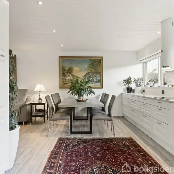 Et moderne køkken med en lang spisebord i midten omgivet af stole. Bordet har en plante omringet af malerier og lamper. Et tæppe ligger på gulvet.