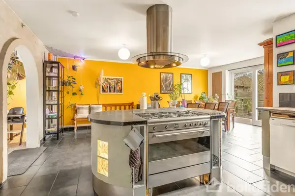 Køkkenø med komfur og emhætte står centralt i et lyst køkken med gule vægge. Rundt om er spiseområde og glasdøre, der åbner mod en have.