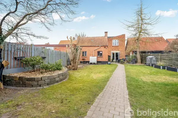 En brostensbelagt sti løber gennem en have mod et murstenshus med rødt tegltag. På begge sider ses græsplæner, træer og buske. En stak af hvide blokke står bag huset.