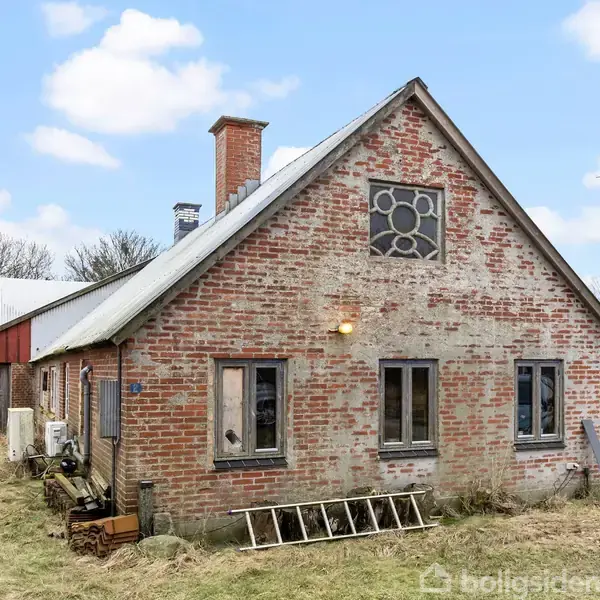 Et rødt murstenshus har et skråt tag og en skorsten, omgivet af græs og træer på en landlig grund med en vindmølle i baggrunden. Murstensfacaden har flere vinduer.