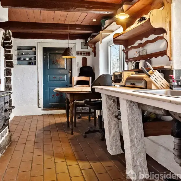 Et landligt køkken med træbjælker og murstensgulv har en keramisk komfur til venstre. Et træbord med stole står i midten, omgivet af åbne hylder fyldt med køkkenudstyr.