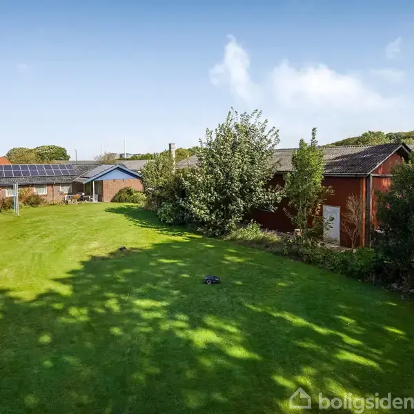 Grønt græsplæneområde med en rød bygning og et hus med solpaneler i baggrunden. Træer og buske grænser op til græsset under en blå himmel.