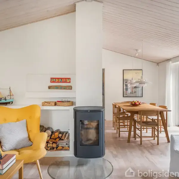 En stue med en gul lænestol ved siden af en bogreol og modelskib. En brændeovn står i midten med brænde under. Et spisebord er i baggrunden ved glasdøre.
