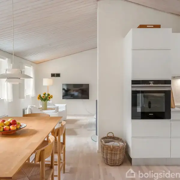 Køkkenalrum med hvidt interiør; spisebord med frugtskål og stole; moderne ovne indbygget i skabsvæg; lysindfald fra vinduer; blomstervase på bord; vægmonteret TV i baggrunden.