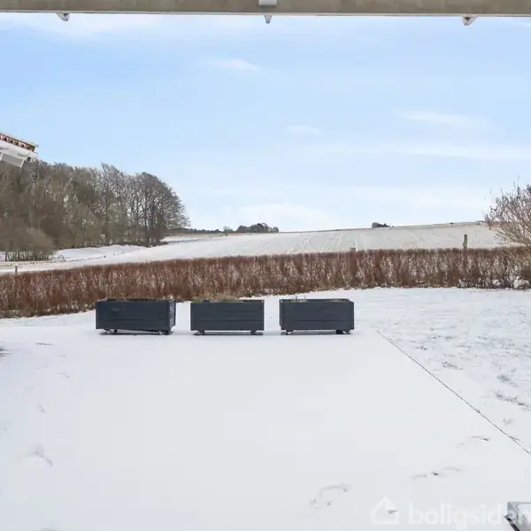 Tre sorte udendørs sofaer står stille på en snedækket terrasse. Omkring er der en hæk, en græsplæne dækket af sne og en klar, blå himmel.