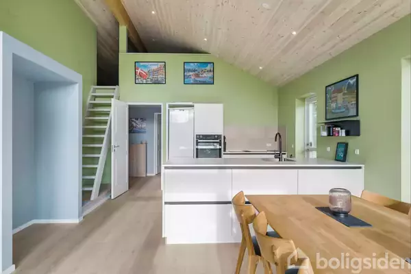 Køkken-alrum med hvid køkkenø og indbyggede apparater står i et rum med lysegrønne vægge og lyst trægulv. Flere malerier hænger på væggene. En trappe fører til en overetage.