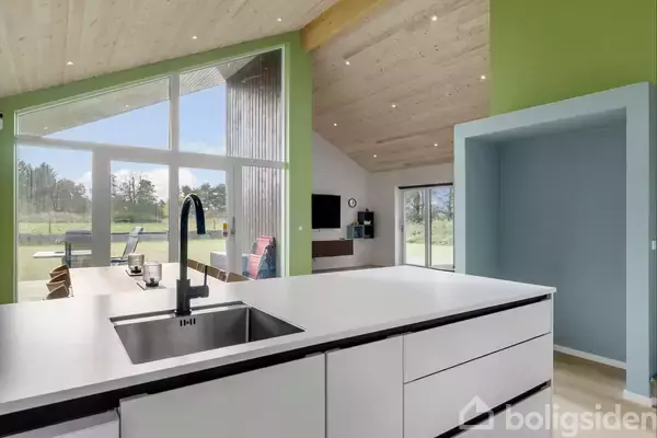 En køkkenø med vask, i en moderne stue med grønne vægge og store vinduer, giver udsigt til en græsplæne og træer udenfor.