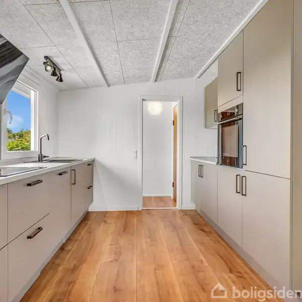 Et moderne køkken med hvide skabe og skuffer langs to vægge. Et vindue og en dør ses for enden af rummet. Trægulv og lyse lofter skaber en lys atmosfære.