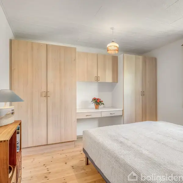 Et soveværelse med en seng placeret ved siden af en trækommode, under en lampe. Garderobeskabe fylder væggen, og der er et vindue med gardiner til højre. En vase med blomster står på en hylde.
