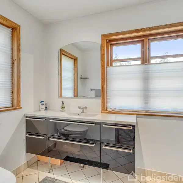 Badeværelse med moderne sort skabsarrangement under spejl, omgivet af hvide vægge og træindrammede vinduer med persienner. Flisegulv og toiletrulleholder ved siden af toilettet.