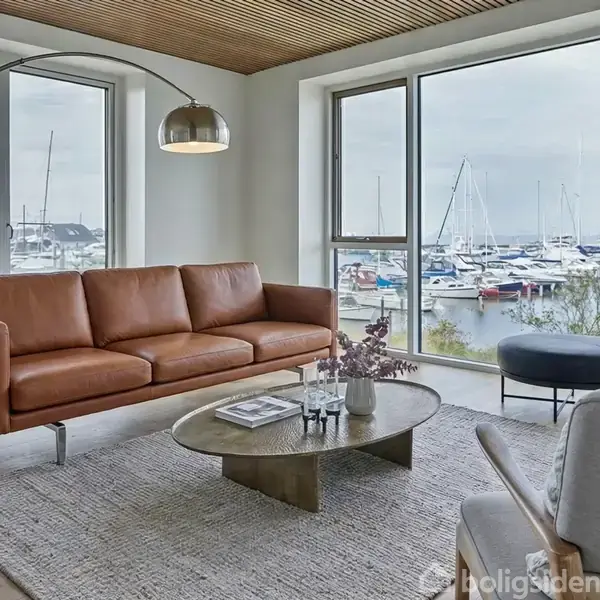 Brun lædersofa står stilfuldt ved et stort vindue med udsigt over en marina. Rundt sofabord med magasiner, flankeret af moderne bue-lampe. Lyst, moderne interiør.