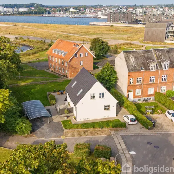 Hus med gråt tag står stille i en boligkvarter. Omgivet af grønne træer og parkeringsareal. Baggrund viser en havnefront med bygninger langs vandet.
