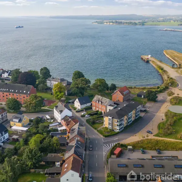 Kystby set fra luften; bygninger langs veje ved vandområde, hvor havet strækker sig ud mod horisonten. Grønt område til højre. Boligsiden vandmærke ignoreret.