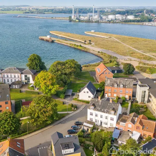 En række huse står stille langs en vej omgivet af grønne træer. I baggrunden ses en kystlinje og et åbent vandområde med industrielt anlæg i horisonten.