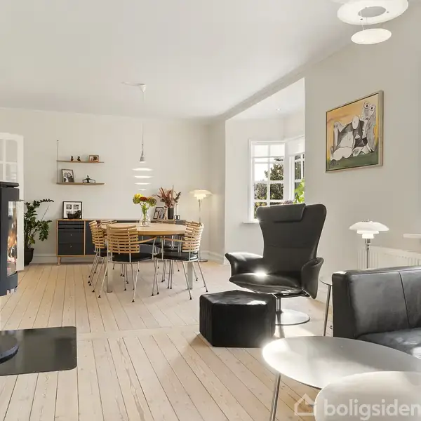 Pejs er tændt i en moderne stue med lyse trægulve. Sofa og lænestol står ved siden af vindue med planter. Spisebord står i baggrunden med pynt og lamper.