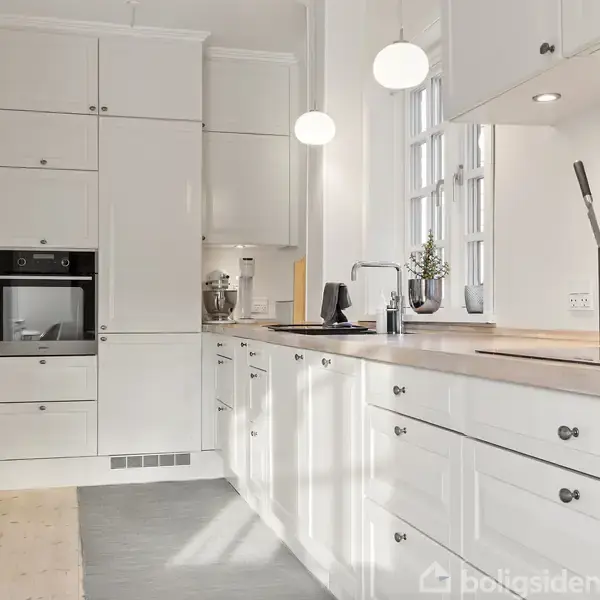 En hvidt, moderne køkken med indbyggede skabe og en sort knivblok, en opvaskemaskine og en indbygget ovn. Rummet er lyst op med runde vedhængslamper og naturligt lys fra vinduerne.