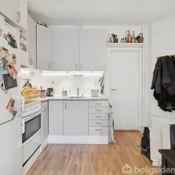 Et køkken med hvide skabe og lysegrå skuffer. En ovn står til venstre, med en garderobehylde til højre med jakker og hjelme. Trægulv dækker gulvet. Tekst på køkkenlåger og køleskab ignoreres.