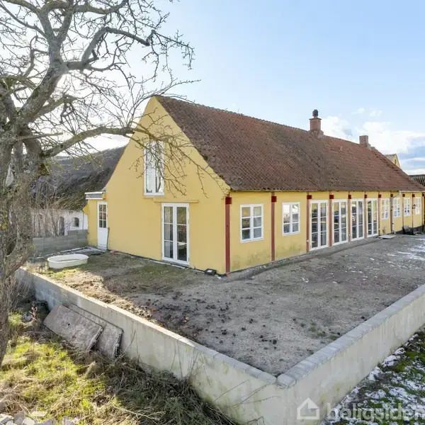 Gul gårdlænge med rødt tegltag står stille i en åben landlig have. Gården har flere vinduer og en terrasse omgives af et træ og åbne marker.