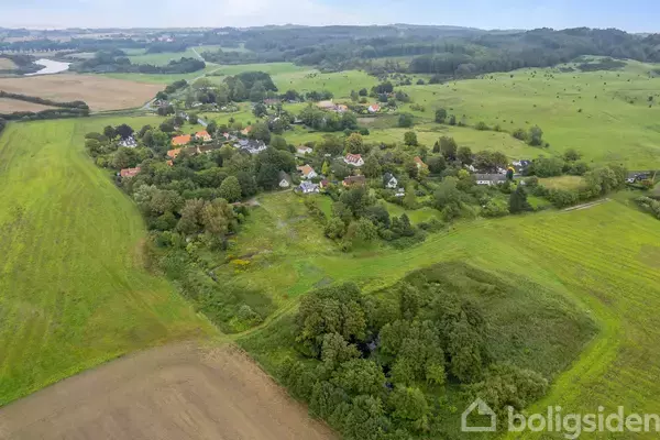 Landsby omgivet af grønne marker og træer, set fra luften. Huse med røde tage spredt blandt træerne, med bakker i baggrunden. Ingen tekst til stede.