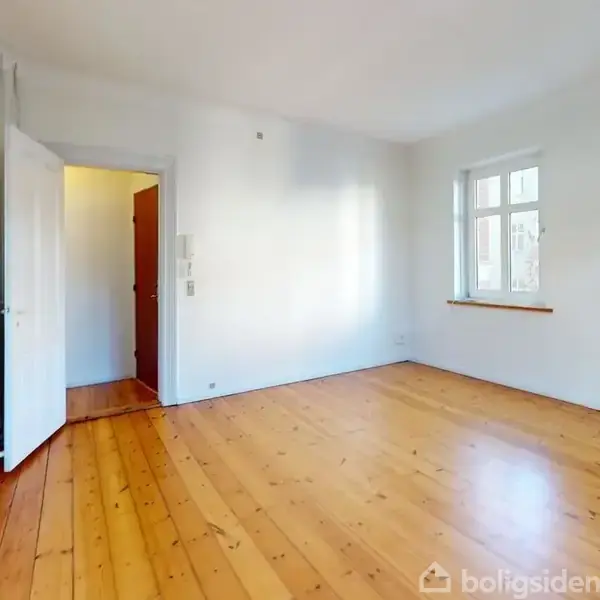 Et tomt værelse med trægulv og hvidmalede vægge. En åben dør fører til en gang. Et enkelt vindue trænger naturligt lys ind.