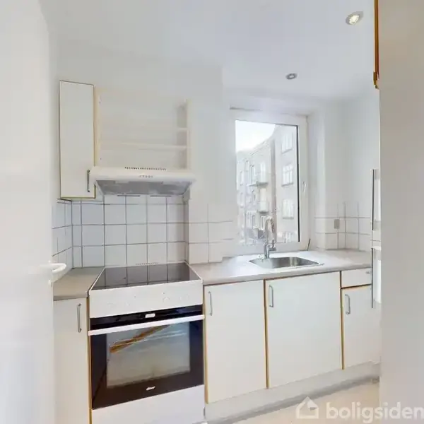 Et lyst køkken med hvide skabe og fliser. En indbygget ovn og vask ses, med et vindue i baggrunden. Fliserne er kvadratiske, og rummet virker rummeligt og veloplyst.