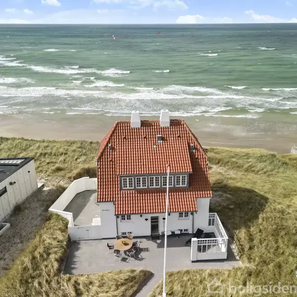 Et rødt hustag med hvide vægge står ved stranden, hvor bølger ruller ind mod kysten. Omgivet af sandklitter, står huset med en terrasse og en parasol i forhaven.