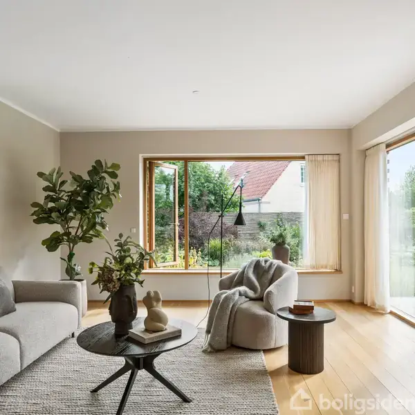 En stue med en lys sofa og et rundt bord prydet med vaser. Store vinduer åbner ud mod en have, og et tæppe ligger på trægulvet.