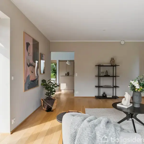 Stue med lys indretning, hvor en sofa står ved et rundt sofabord med blomster. En reol med dekorationer ses mod væggen, og store vinduer lader sollys komme ind.