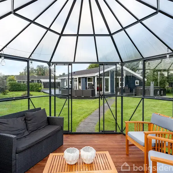 En møbleret havepavillon med glasvægge står i en grøn have. Sort sofa og træstole omgiver et bord med dekorative lys. I baggrunden ses et moderne hus.