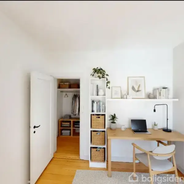 Et skrivebord står i et lyst rum med en åben dør til en garderobe. Skrivebordet har en lampe, bærbar computer, planter og bøger. Hylder og kurve supplerer den minimalistiske indretning.
