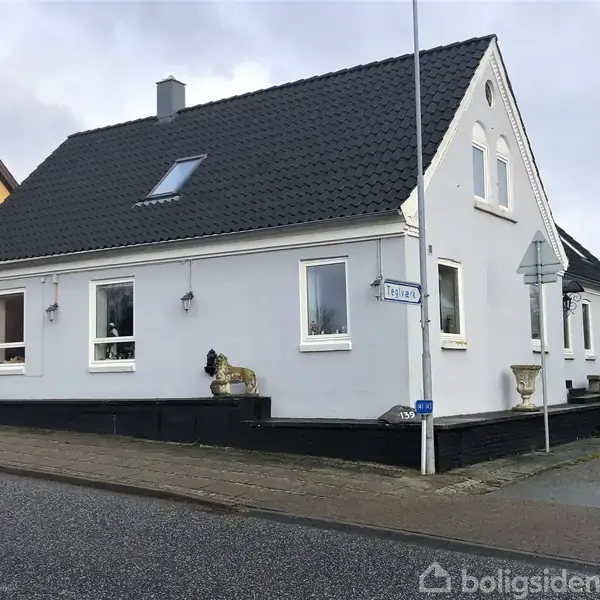 Et hvidt hus med sort tag står på hjørnet af en gade, ved siden af fortovet. Skiltet viser "Løgstrupvej 139". Flere vinduer vender mod gaden.