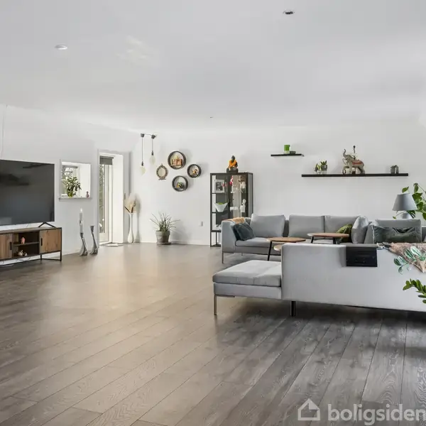 Stue med stor sofa og TV på skænk. Indretning inkluderer planter, væghylder med dekoration, og lysindfald fra vinduer i moderne og rummeligt interiør.