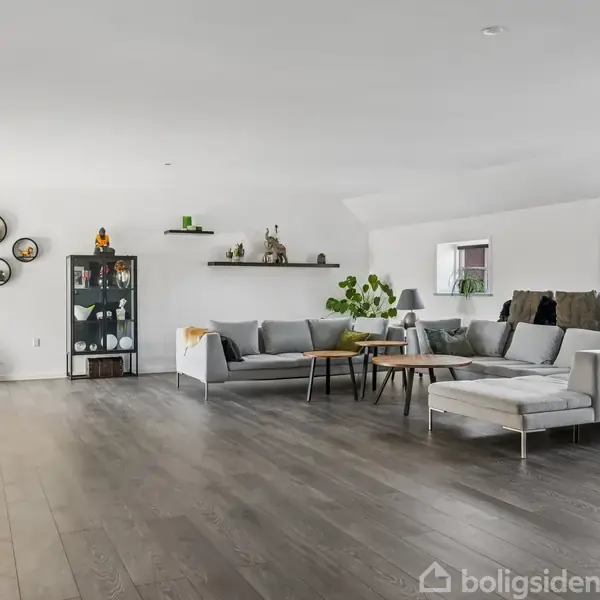 Stor stue med en grå hjørnesofa, sofabord og grønne planter. Baggrunden har vægrammer og hylder med pyntegenstande. Trægulv og tre små vinduer skaber en lys, luftig stemning.