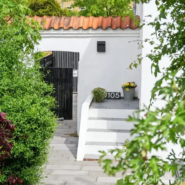 Hvid murstensvæg med trin op til hoveddør, nummer 105 synligt. Omkringliggende grønne buske og planter skaber en frodig haveatmosfære.