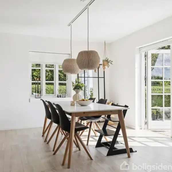 Spisebord med skåle og lamper hængende over i en lys spisestue. Seks stole omkring bordet. Store vinduer og glasdøre åbner ud mod en grøn have.