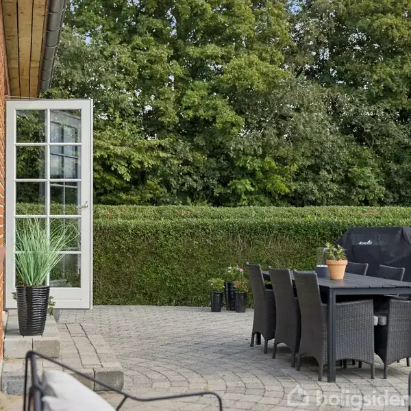 En åben terrassedør fører ud til en solrig terrasse med sorte havemøbler og potteplanter. I baggrunden er der en veltrimmet hæk og grønne træer.
